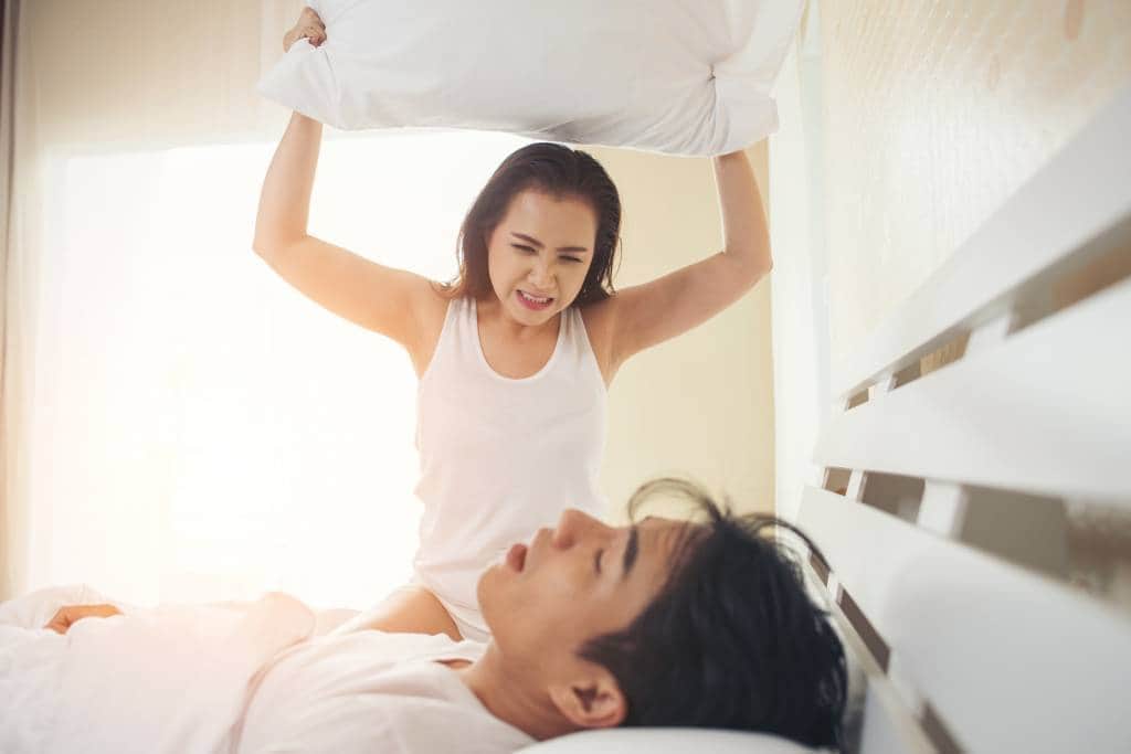 woman holding pillow above snoring partner in bed
